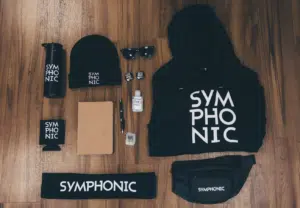 A flat lay of "SYMPHONIC" branded merchandise on wood floor, including hoodie, fanny pack, beanie, tumbler, koozie, notebook, headband, sunglasses, pens, and hand sanitizer—perfect essentials for the Amsterdam Dance Event.